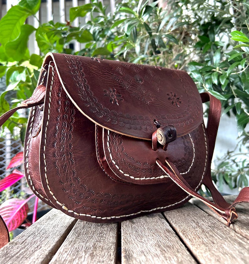 May include: A brown leather saddlebag with floral embossed detailing and white stitching. The bag features a flap closure with a wooden toggle and a long adjustable strap. The bag is sitting on a wooden surface.