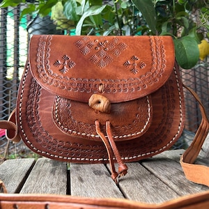 May include: Brown leather crossbody bag with embossed floral and geometric patterns. The bag features a flap closure with a wooden toggle and a long adjustable strap. White stitching accents the edges of the bag.