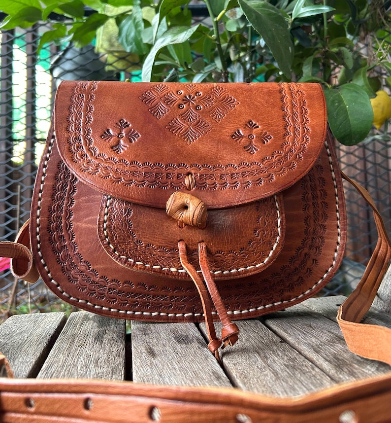 May include: Brown leather crossbody bag with embossed floral and geometric patterns. The bag features a flap closure with a wooden toggle and a long adjustable strap. White stitching accents the edges of the bag.