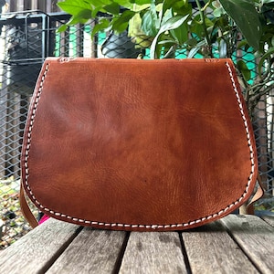 May include: A brown leather saddlebag with a curved bottom and a flap closure. The bag features white stitching along the edges and a long strap. The bag is made of a textured leather material.