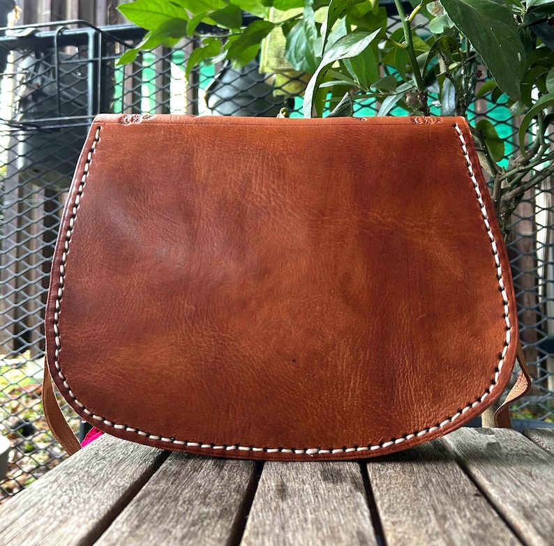 May include: A brown leather saddlebag with a curved bottom and a flap closure. The bag features white stitching along the edges and a long strap. The bag is made of a textured leather material.
