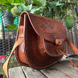 May include: A brown leather crossbody bag with a flap closure and a long adjustable strap. The bag features intricate embossed floral patterns and white stitching along the edges. The bag is made of genuine leather.