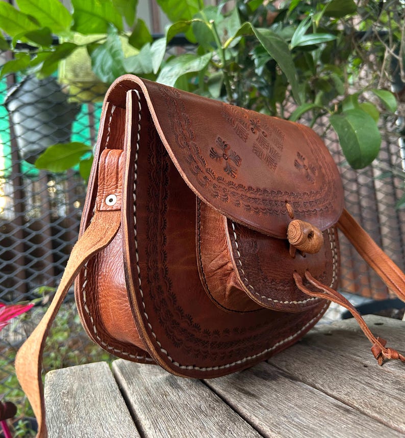 May include: A brown leather crossbody bag with a flap closure and a long adjustable strap. The bag features intricate embossed floral patterns and white stitching along the edges. The bag is made of genuine leather.