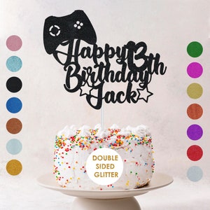 May include: Black glitter cake topper featuring a video game controller, the words "Happy 13th Birthday Jack" and two stars. The topper is on a white cake with colourful sprinkles. The cake is on a white cake stand. A label on the cake says "Double Sided Glitter".