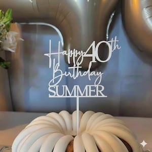 May include: A white cake topper with the words "Happy 40th Birthday SUMMER" in a script font. The topper is inserted into a white frosted cake. The background includes silver balloons and a bouquet of white flowers.