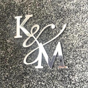 May include: Silver acrylic letters 'K' and 'M' in a decorative script. The 'K' is a standard capital letter, while the 'M' is a capital letter with a modern font. The letters are intertwined with a cursive flourish.