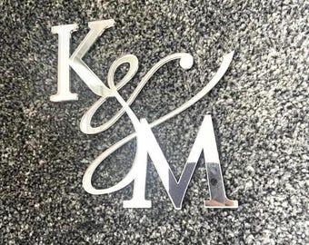 May include: Silver monogram with the letters "K" and "M" in a decorative script. The letters are made from a reflective material, giving a mirrored appearance. The monogram is set against a textured, grey background.
