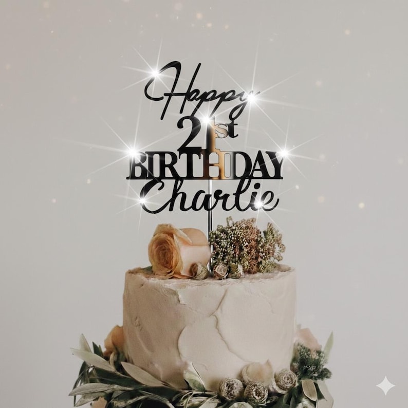 K&ouml;nnte beinhalten: Eine Geburtstagstorte mit einem schwarzen Acryl-Kuchenaufsatz mit der Aufschrift "Happy 21st Birthday Charlie". Die Torte ist mit Blumen und Gr&uuml;n geschm&uuml;ckt. Die Torte hat ein wei&szlig;es Frosting.