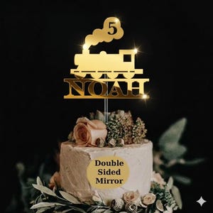 May include: A gold-coloured cake topper shaped like a train, featuring the number 5 and the name "NOAH". The topper sits atop a white frosted cake adorned with flowers and foliage. The words "Double Sided Mirror" are also visible on the cake.