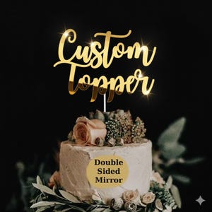 May include: A gold-coloured 'Custom Topper' cake decoration with a reflective surface, placed atop a white frosted cake adorned with flowers and greenery. The cake has a 'Double Sided Mirror' label.