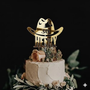 May include: A gold-coloured cake topper shaped like a cowboy hat, displaying the number 30 and the name "HENRY". The topper sits atop a white frosted cake, adorned with flowers and foliage. The backdrop is black.
