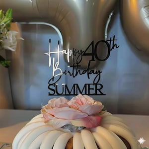 May include: A black and silver cake topper that reads "Happy 40th Birthday SUMMER". The topper is placed on top of a white and brown cake decorated with a pink flower.