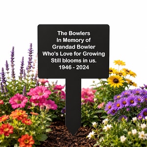 May include: A black rectangular garden stake with a pointed bottom, featuring white text that reads: The Bowlers, In Memory of, Grandad Bowler, Who's Love for Growing, Still blooms in us., 1946 - 2024. The stake is positioned among colourful flowers.