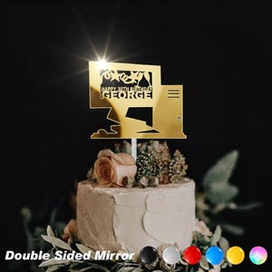 May include: A gold mirror cake topper with the text "HAPPY 30TH BIRTHDAY GEORGE". The topper is in the shape of a computer monitor and is placed on top of a white frosted cake decorated with flowers and greenery.