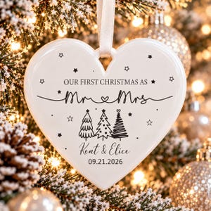May include: A white heart-shaped ceramic ornament hanging from a white ribbon. The text reads "OUR FIRST CHRISTMAS AS Mr & Mrs", "Kent & Elice", and "09.21.2026". It features black line art of three stylized Christmas trees and small stars.