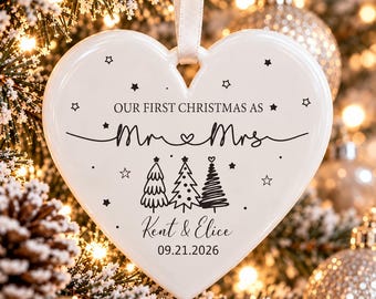 May include: A white heart-shaped ceramic ornament hanging from a white ribbon. The text reads "OUR FIRST CHRISTMAS AS Mr & Mrs", "Kent & Elice", and "09.21.2026". It features black line art of three stylised Christmas trees and small stars.