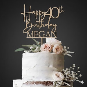 May include: A two-tiered white cake decorated with flowers and a wooden cake topper. The topper reads "Happy 40th Birthday MEGAN" in a script font. The cake is adorned with pink and white roses and greenery.