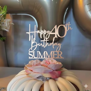 May include: A rose gold cake topper that reads "Happy 40th Birthday SUMMER" is placed on top of a frosted cake with a pink flower. The cake is decorated with white frosting and sits on a white surface. The background includes a blurred silver balloon.