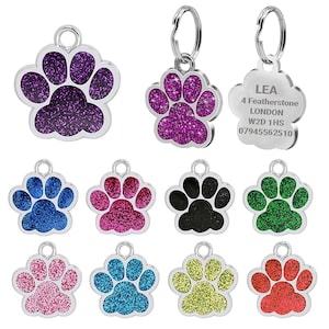 May include: A selection of paw print-shaped pet ID tags in various colours, including purple, blue, pink, black, green, yellow, and red, with a glitter finish. One tag is engraved with the name "LEA" and contact details.