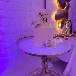 May include: A two-tiered wedding cake with white icing, decorated with flowers and gold initials. The cake is on a round table with a white top and gold base, creating an elegant display. The table is approximately 76 cm in diameter.