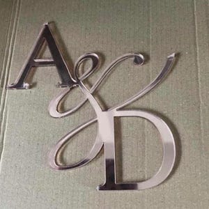 May include: Silver metal letters 'A', 'K', and 'D' in a decorative script font. The letters are placed on a textured, light green surface. The 'K' and 'D' are connected, while the 'A' is separate.