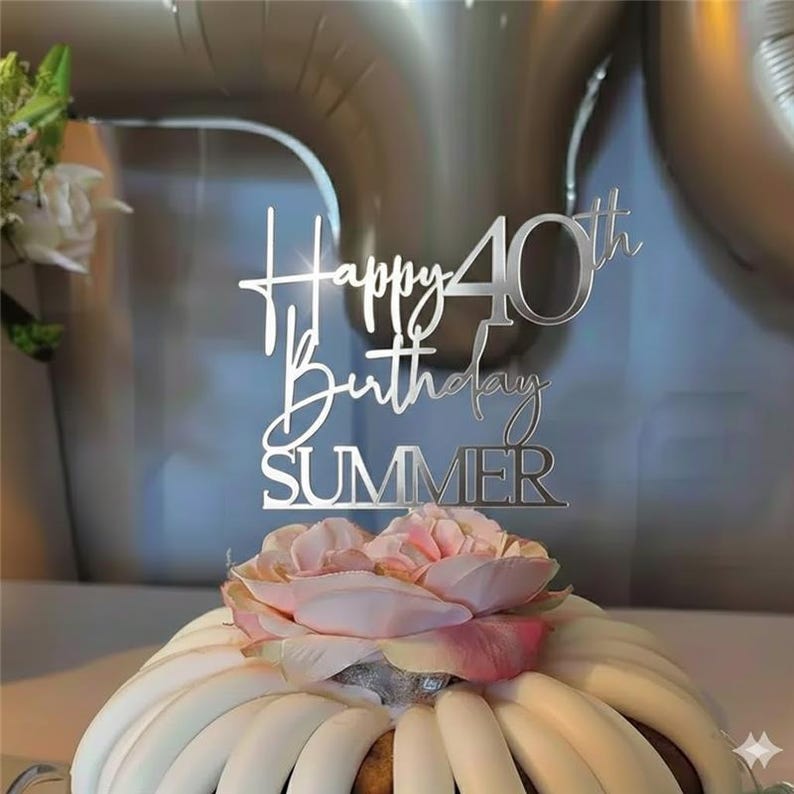 May include: A silver cake topper with the words "Happy 40th Birthday SUMMER" is placed on top of a cake decorated with white frosting and a pink flower. The background includes silver balloons.