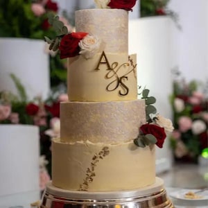 May include: A four-tiered wedding cake with a cream base and textured layers. The cake is decorated with red and white roses, gold initials, and gold leaf accents. It sits on a silver cake stand.