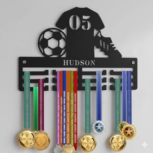 May include: A black metal wall-mounted medal hanger featuring a soccer jersey silhouette with the number 05, a soccer ball, and a cleat. The horizontal bar displays the name HUDSON and includes multiple hooks holding various colorful ribbons with medals.