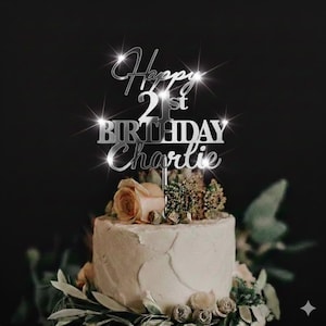 K&ouml;nnte beinhalten: Ein wei&szlig;er Kuchen mit Blumendekorationen, gekr&ouml;nt von einem silbernen Tortenaufsatz mit der Aufschrift "Happy 21st Birthday Charlie". Der Kuchen steht vor einem dunklen Hintergrund.