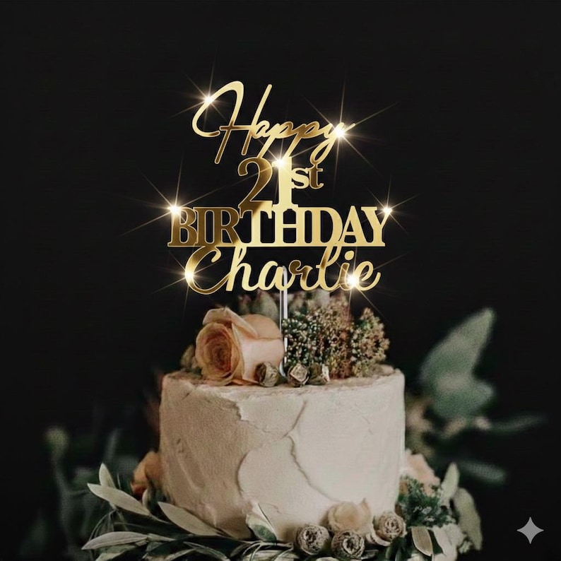 K&ouml;nnte beinhalten: Ein goldfarbener Tortenaufsatz mit der Aufschrift "Happy 21st Birthday Charlie" befindet sich auf einem wei&szlig; glasierten Kuchen, der mit Blumen und Gr&uuml;n geschm&uuml;ckt ist. Der Text hat einen Glitzereffekt vor dunklem Hintergrund.