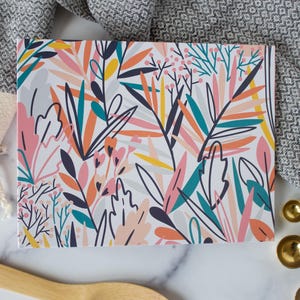 May include: A rectangular notebook with a colorful floral design featuring pink, orange, teal, and yellow leaves. The notebook is set on a white surface with a gray patterned towel, wooden spoon, and gold measuring spoons.