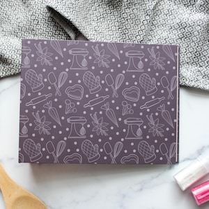May include: A rectangular box with a purple background and a white pattern of baking tools, including mixers, rolling pins, and oven mitts. The box is surrounded by a wooden spoon, a gray patterned cloth, and two small bottles of sprinkles.