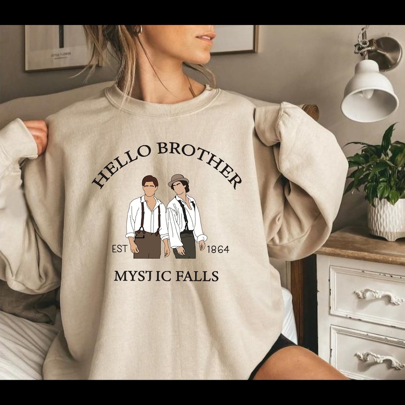 May include: A beige sweatshirt with the text "HELLO BROTHER EST 1864 MYSTIC FALLS" in black. The sweatshirt features a graphic of two men in black and white.