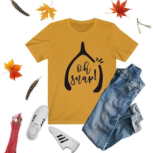 May include: A mustard yellow t-shirt with a black graphic of a wishbone and the text "Oh snap!"  The t-shirt is lying on a white background with a pair of white trainers, a pair of blue jeans, and some autumn leaves.
