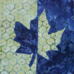 May include: A blue fabric leaf with a jagged edge is sewn onto a green fabric background with a pattern of small circles. The leaf is partially overlapping the edge of the fabric.