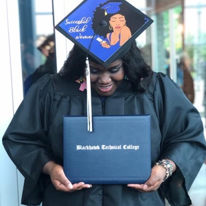 May include: A graduate in a black gown and cap, holding a diploma from Blackhawk Technical College. The cap is decorated with a blue and black design featuring the text "Successful Black Woman" and an illustration of a woman.
