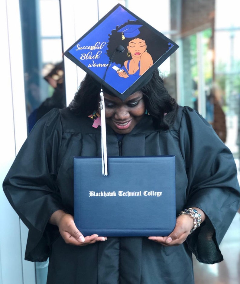 May include: A graduate in a black gown and cap, holding a diploma from Blackhawk Technical College. The cap is decorated with a blue and black design featuring the text "Successful Black Woman" and an illustration of a woman.