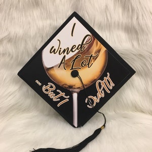 May include: Black graduation cap with the text "I Wined A Lot... But I Did It!" and a wine glass graphic. The cap has a black tassel with a gold accent.