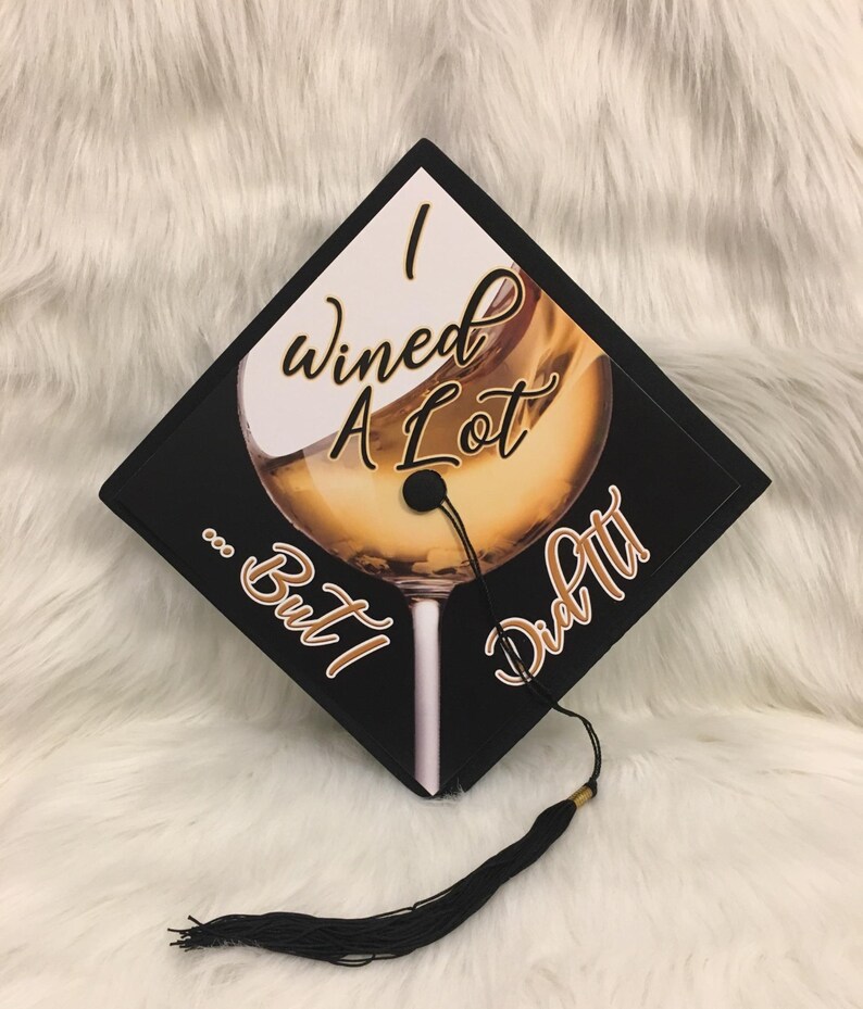 May include: Black graduation cap with the text "I Wined A Lot... But I Did It!" and a wine glass graphic. The cap has a black tassel with a gold accent.