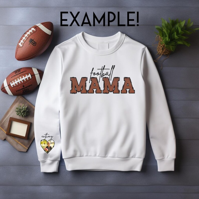 Może przedstawiać: Biała bluza z brązowym napisem "football mama". Bluza ma małe serce na rękawie z włożonym zdjęciem.