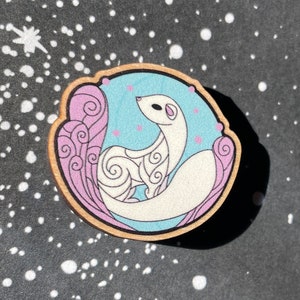 May include: Wooden pin featuring a white weasel with a patterned design, set against a light blue background with pink and purple accents. The pin is round with a natural wood border, and is set against a black background with white dots.