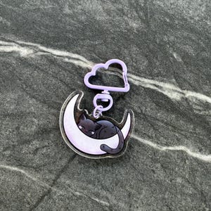 May include: A clear acrylic keychain featuring a black cat sleeping on a light purple crescent moon. The charm is attached to a light purple cloud-shaped clasp. The design includes a dark, glittery background behind the cat and moon.
