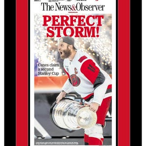 May include: A framed newspaper front page titled The News & Observer with the headline PERFECT STORM! featuring a hockey player in a white, red, and black jersey holding the Stanley Cup trophy. The frame has a black outer border and a red inner mat.