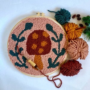 mushroom needle punch in a large embroidery hoop with green laurel leaves. Brown, yellow, green and pink yarn next to it. Needle punch with yarn sits on top of it.