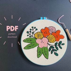Op de afbeelding: Een blauwe borduurring met een witte stoffen achtergrond. De stof is versierd met een kleurrijk bloemenontwerp gemaakt met de punch needle-techniek. Het ontwerp bevat oranje, gele, roze en groene bloemen. De tekst "PDF pattern download" is zichtbaar in de linkerbovenhoek van de afbeelding.