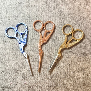 May include: Three pairs of decorative scissors with bird-shaped handles. One pair is blue and white, one is rose gold, and one is gold.