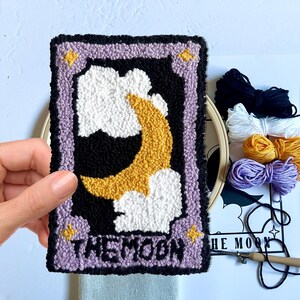May include: A purple and black needlepoint embroidery of a crescent moon with clouds and stars. The embroidery is framed with a lavender border and the text "THE MOON" is stitched in black thread.