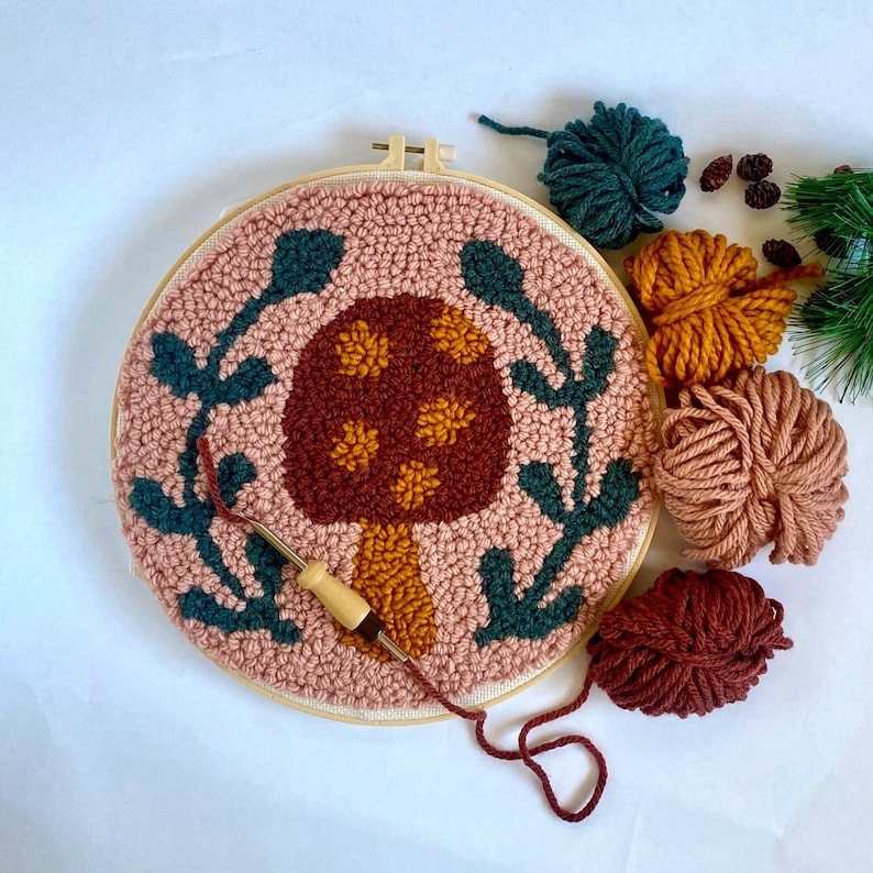 mushroom needle punch in a large embroidery hoop with green laurel leaves. Brown, yellow, green and pink yarn next to it. Needle punch with yarn sits on top of it.