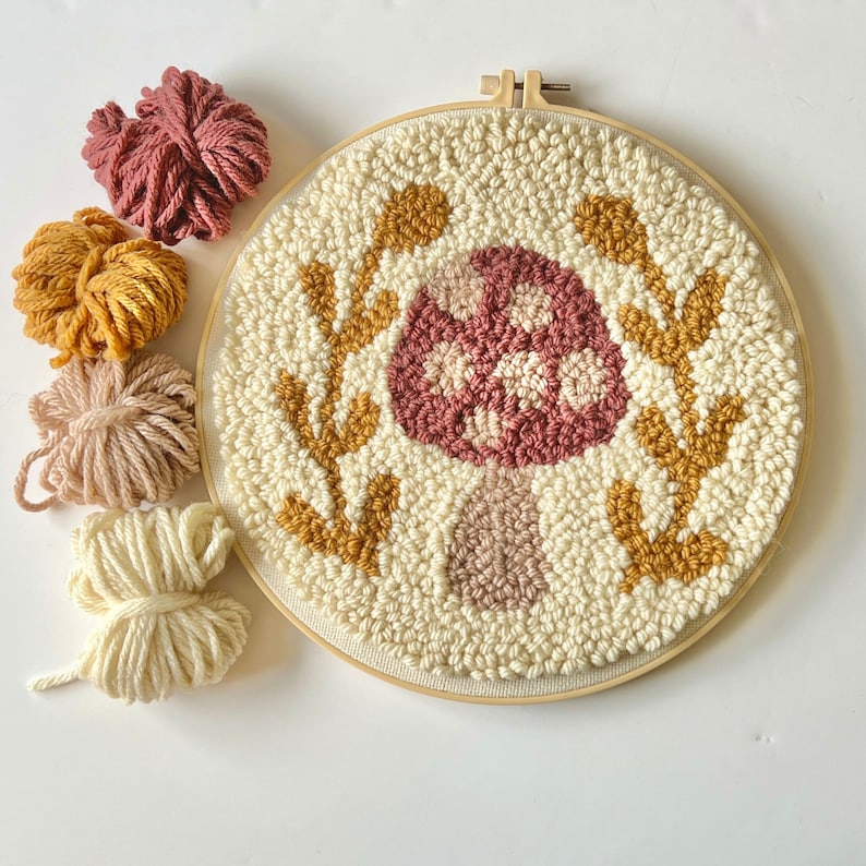 mushroom needle punch in a large embroidery hoop with yellow laurel leaves. Pink, beige, yellow, white yarn next to it.