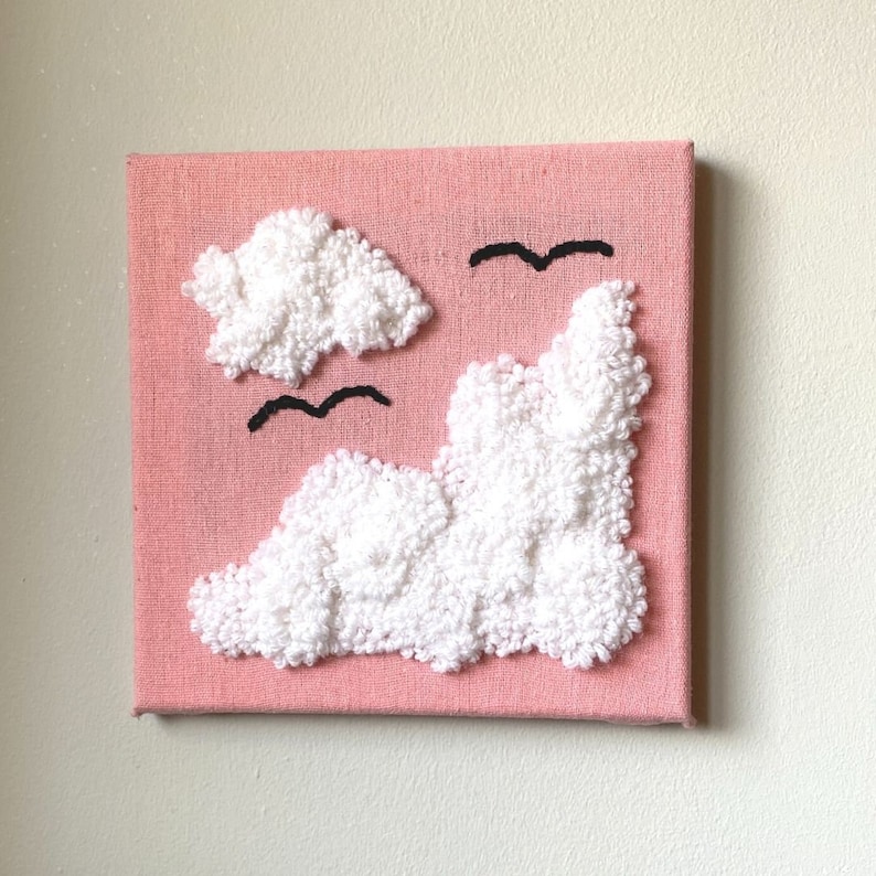 Square frame with pink cotton linen stretched over it with white sparkling clouds made with punched yarn and 2 black yarn seagulls. Wall art.